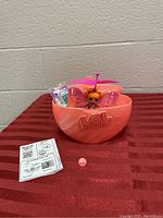 LOL Surprise Magic Flyers doll with wings and propeller inside pink carrying case on red surface. Instruction manual and small pink round accessory visible next to the case.