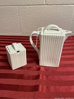White ceramic tea pot and sugar dish set placed on a red striped fabric surface, showing side angles and design details