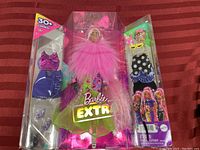 Barbie Extra doll in original pink and glittery packaging, showing doll wearing large pink feathered garment and multicolor outfit.