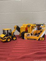 Photo showing the yellow and black CAT toy excavator and the boxed CAT dump truck sand set side by side on a red surface.