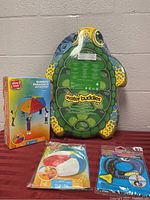 Photo showing three items on table: large turtle-shaped Water Buddies foam float board, boxed Rainbow Parachute Activity set, packaged inflatable beach ball and blue diving toy.