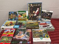 Complete lot of 12 assorted jigsaw puzzles displayed on table. Boxes show different nature, animal and holiday scenes.
