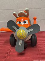 Front view showing orange airplane-themed ride-on toy with large spinning propeller, eyes, and antennae.