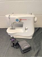 Singer M2100 sewing machine with foot pedal and power cord placed on black surface in front of cinder block wall