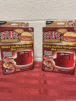 Two boxed Stufz burger makers side by side on red cloth background, both with packaging intact, one with plastic wrap, one without.