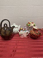 Photo showing full view of the 5 teapots arranged on a red cloth, showing details and colors of each teapot.