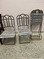 Four vintage chairs with cast iron frames and backs, wooden slatted seats with faded blue/gray paint, two stacked and two standing upright on terrazzo flooring.