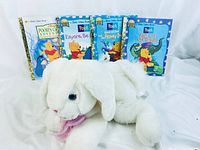 Photo showing the four Winnie the Pooh Little Golden Books standing upright, with the white plush rabbit lying in front of them. Books have illustrated colorful covers and gold spines. Rabbit is white with floppy ears and a pink ribbon tied at the neck.