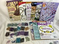 Multiple sealed small bags of glass pearl beads laid out by color and size in front of 5 different beading/jewelry books and magazines.
