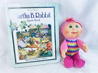 Photo showing Martha B. Rabbit Jigsaw Book standing beside the Cabbage Patch Kid doll on a white background. The book cover has visible wear around the edges.