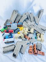 Wide view showing five different colored Thomas the Train engines, a brown wagon, many gray plastic train track pieces bundled together, and station house with bridge supports.