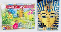 Boxed 1000-piece Art Gallery jigsaw puzzle showing an outdoor garden scene with flowers, birdhouse, and small bridge, alongside the Tutankhamun puzzle book with the golden mask cover.