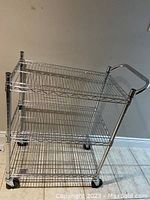 Front view of three-tier chrome-plated wire storage cart on swivel casters against wall.