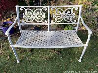 Front view of white metal 2-seater bench with lattice seat and scroll backrest, outdoor on grass with plants in background.