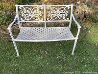 Full view of white metal outdoor bench with floral scroll pattern on backrest and lattice seat on artificial grass