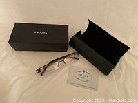 Prada glasses frames alongside black hard case, Prada branded box and authenticity certificate card.