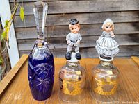 Set of three decorative bells displayed outdoors on wooden surface against wooden wall. Left bell is blue cut glass with clear faceted handle, right two are clear glass with gold accents and porcelain figurine handles.