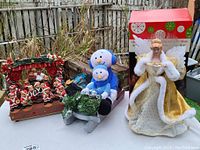 Shows all three pieces: bear music band figurine on a stage, plush snowmen on sled, angel tree topper doll with box in background