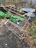 Wooden folding clothes drying rack positioned outdoors on a leaf-covered ground with miscellaneous background items.