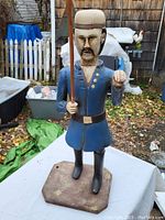 Front view of carved folk art figure standing on a flat base, dressed in blue coat with gold buttons, black boots, holding ball in left hand and scythe in right hand.