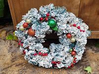 Front view of a large flocked winter wreath with red berry clusters and multi-color glittery ornaments hanging on a wooden background.
