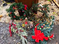 Five assorted Christmas wreaths laid on the ground with various decorations including ribbons, berries, pine cones, and a large red bow.