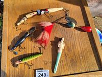 Photo showing all seven vintage fishing lures laid out on a wooden surface in sunlight, displaying colours and details of each item in the lot.
