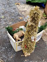 Photo showing a tall gold tinsel Christmas tree with mixed colored ornaments next to a box containing a green garland and a brown wood-like reindeer figure