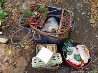 Wide shot showing the wicker sleigh with greenery and berries, plastic bin with assorted decor, and a ceramic Santa Claus container among other holiday items.