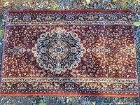 Photo of small rectangular Turkish rug with detailed red, blue, ivory geometric and floral patterns, shown on ground outdoors.