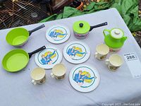 Vintage 1983 Barbie dishes and kitchenware lot including 5 round plates featuring Barbie and Ken, 4 mugs with Barbie prints, and 3 cooking pieces in bright green with black handles.