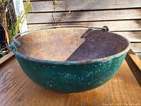 Photo showing the vintage cast iron bean pot with green exterior paint and rusted interior, placed on a wooden surface outdoors.