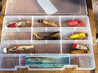 Top view of plastic tackle box showing vintage fishing lures of different shapes, sizes, and colors arranged in compartments.