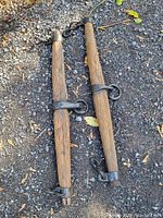 Two wooden whiffletrees with blacksmith made metal rings and hooks, placed on gravel ground.