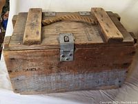Front angled view of vintage wooden military box showing rope handle, wooden slats on lid, metal latch, and stencil markings.