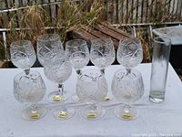 Set of 9 clear glass wine glasses with etched cornflower floral pattern arranged in two rows on a white cloth, outdoor setting.