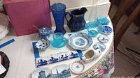 Photo showing the full lot including two blue glass vases, a blue glass pitcher, blue glass saucers, a sugar bowl, glass shoe figurine, a variety of decorative mini plates with blue scenic and floral patterns.