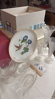 Photo of glassware including vases, punch bowl, and hummingbird plate on table with boxes in background