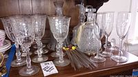 Clear etched floral glass goblets, cut crystal decanters, glass stir sticks, and commemorative flutes displayed on wooden surface