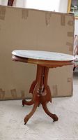 Vintage oval marble top table with wood pedestal base in front of a large cardboard backdrop. Marble top oval shape light colored with some veining visible. Wood base with curved legs ending in small casters.