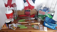 Photo of the lot including grill tools with green handles, red and white cooler, drinking glasses, Putting Partner golf aid, blue pouch, beer opener, and flower seed packets on a table.