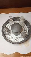 Metal tray holding a glass perfume bottle with faceted clear stopper, another with frosted stopper, a third with a round stopper, a round metal powder canister, and a small metal container with engraved lid on a doily.