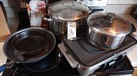 Photo showing large and medium stainless steel stock pots with lids, small cast iron pan, Cuisinart electric griddle, and Vslicer tool on stove