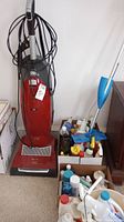 Miele red and gray upright vacuum cleaner with black power cord wrapped around handle and flexible hose, next to boxes containing assorted cleaning products including sprays and liquids.