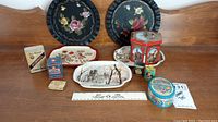 A full view of all items in the lot showing two round hand painted black metal plates with floral motifs, multiple small vintage tins including a Colgate baby talc tin, a red Lowney's cocoa tin, small decorative trays with scenic prints, and other small tins.