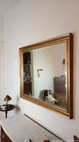Frontal view of rectangular wall mirror with beveled edge and gold-colored wide frame mounted on a wall, showing reflection of room interior.