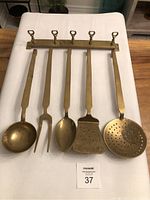 Front view of six brass kitchen utensils hanging from a brass wall rack with six hooks, laid flat on a white cloth surface.