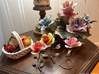 Four Capo di Monte porcelain pieces including three roses on metal stems and one fruit basket with fruit and flowers on a wooden surface.