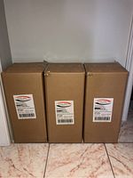 Three unopened boxes containing pendant light fixtures by Dolan Hospitality stacked side by side on a tiled floor.