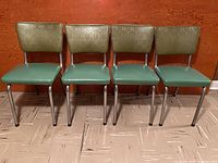 Front view of four retro kitchen chairs in a row with green vinyl seats and patterned green vinyl backrests against an orange wooden wall